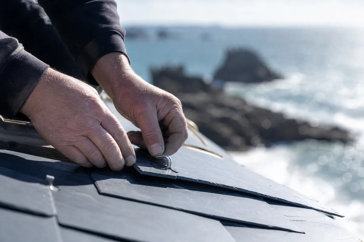 Mains d’un couvreur fixant un crochet inox sur une ardoise, mer d’Iroise en arrière-plan